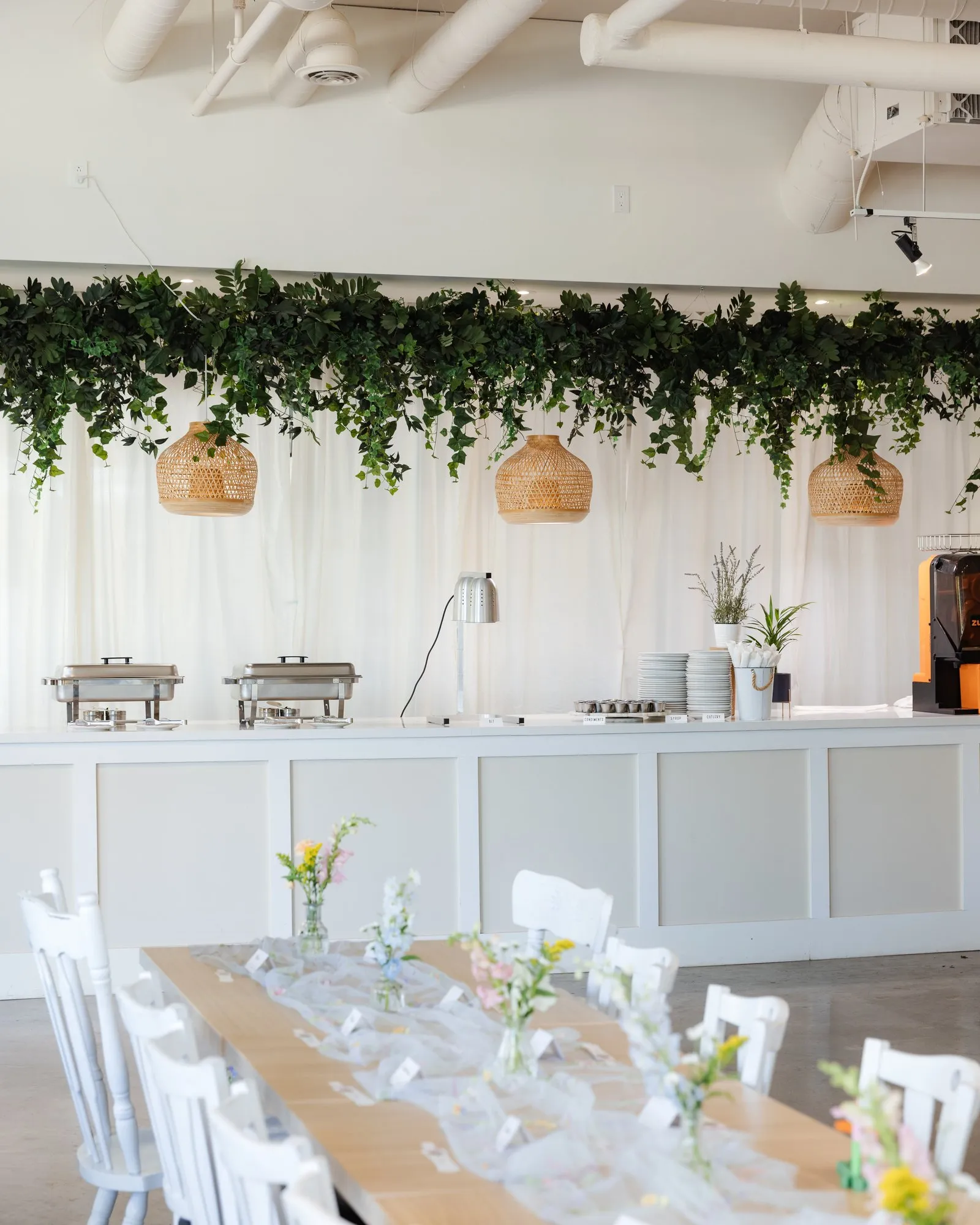 Long table with a tulle runner and wildflower bud vases under a hanging greenery garland and rattan pendant lights