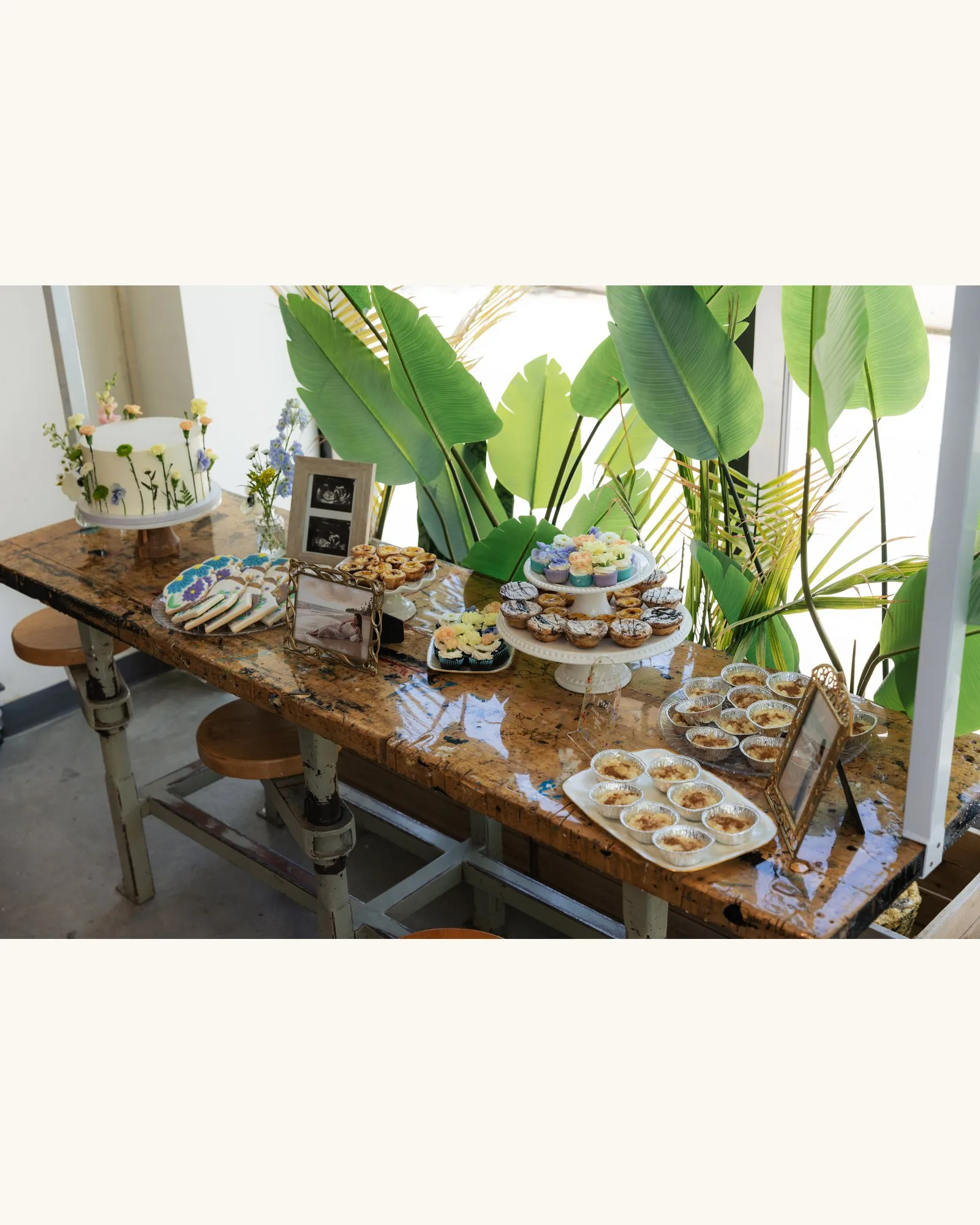 Dessert table on a rustic wooden bench against tall tropical leaves, with the cake, cookies, cupcakes and tarts