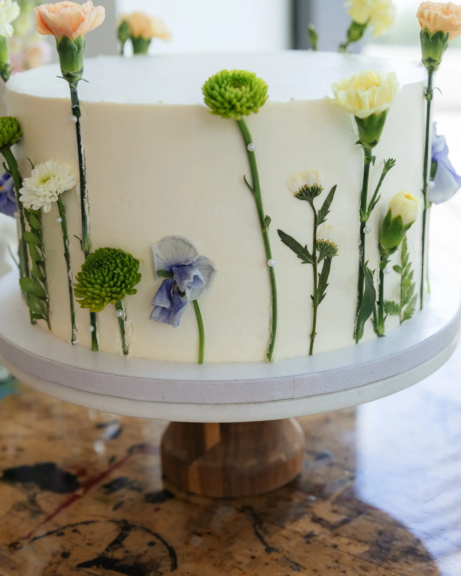 White cake decorated with pressed carnations, chrysanthemums and violas on a wooden cake stand