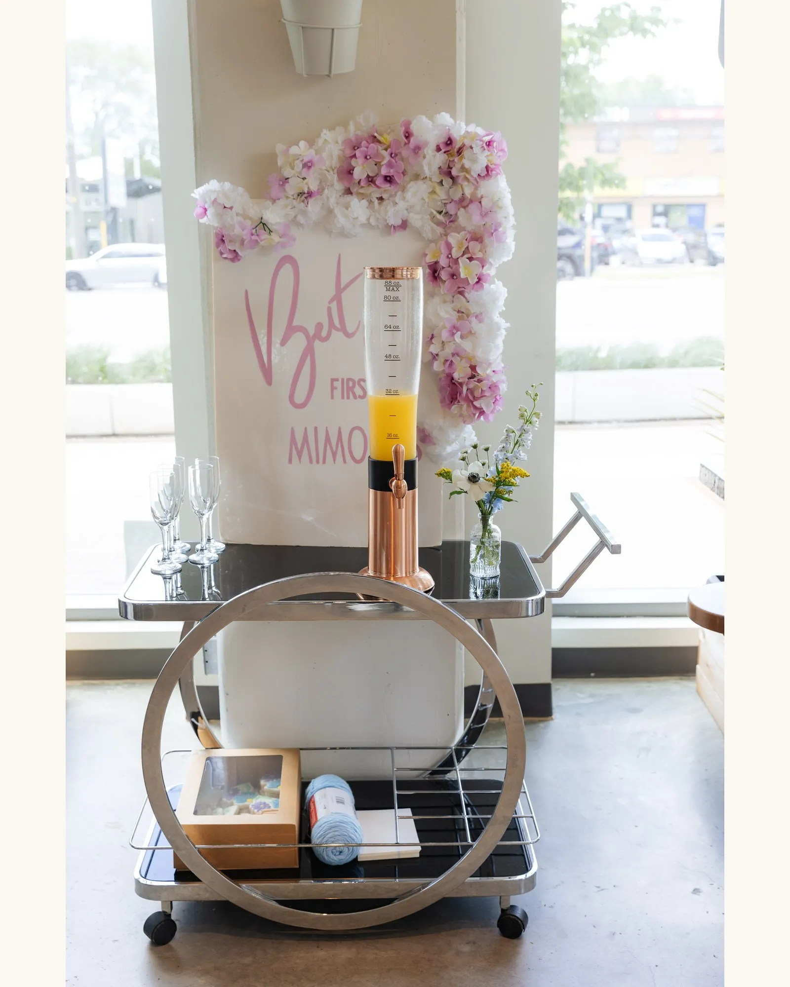 Mimosa bar cart with a juice tower and glasses under a pink and white flower arch and a "But first mimosas" sign